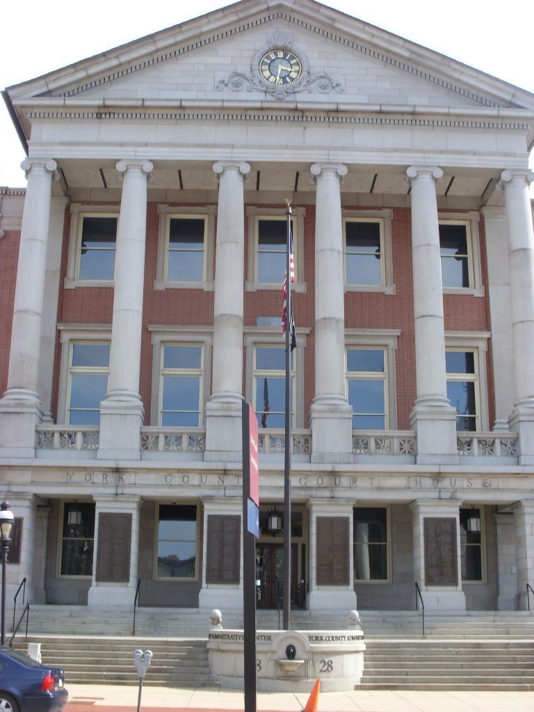 Two-story brick building with white columns and clock, likely a courthouse, with flags and a car.