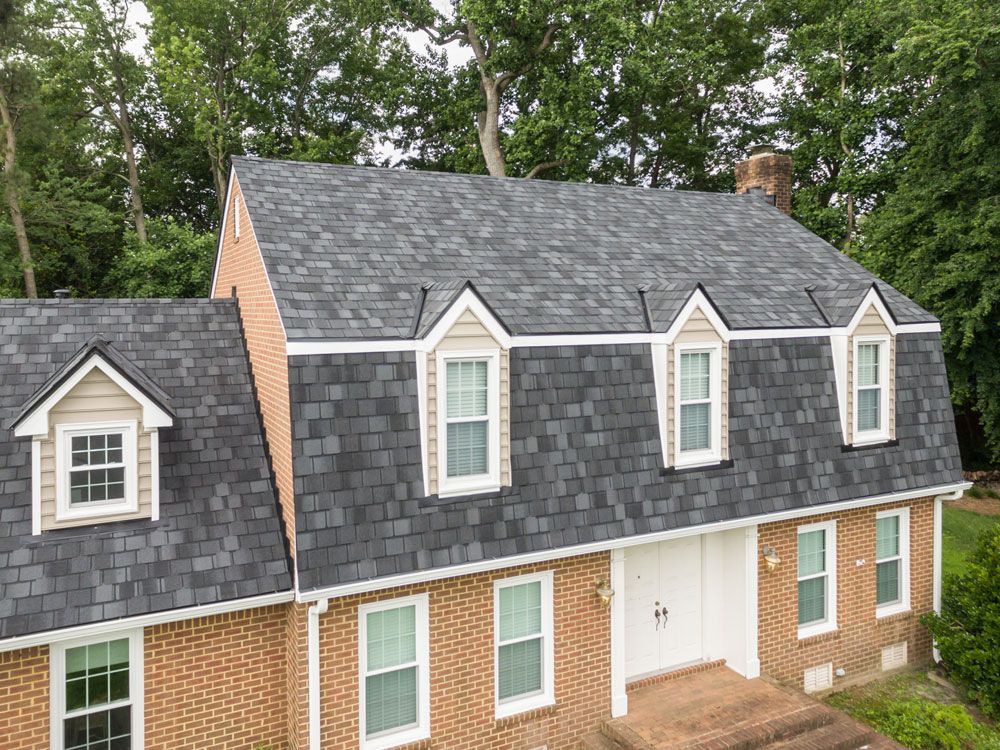 Gray shingle roof on a two-story brick house with white trim and dormer windows, surrounded by trees.