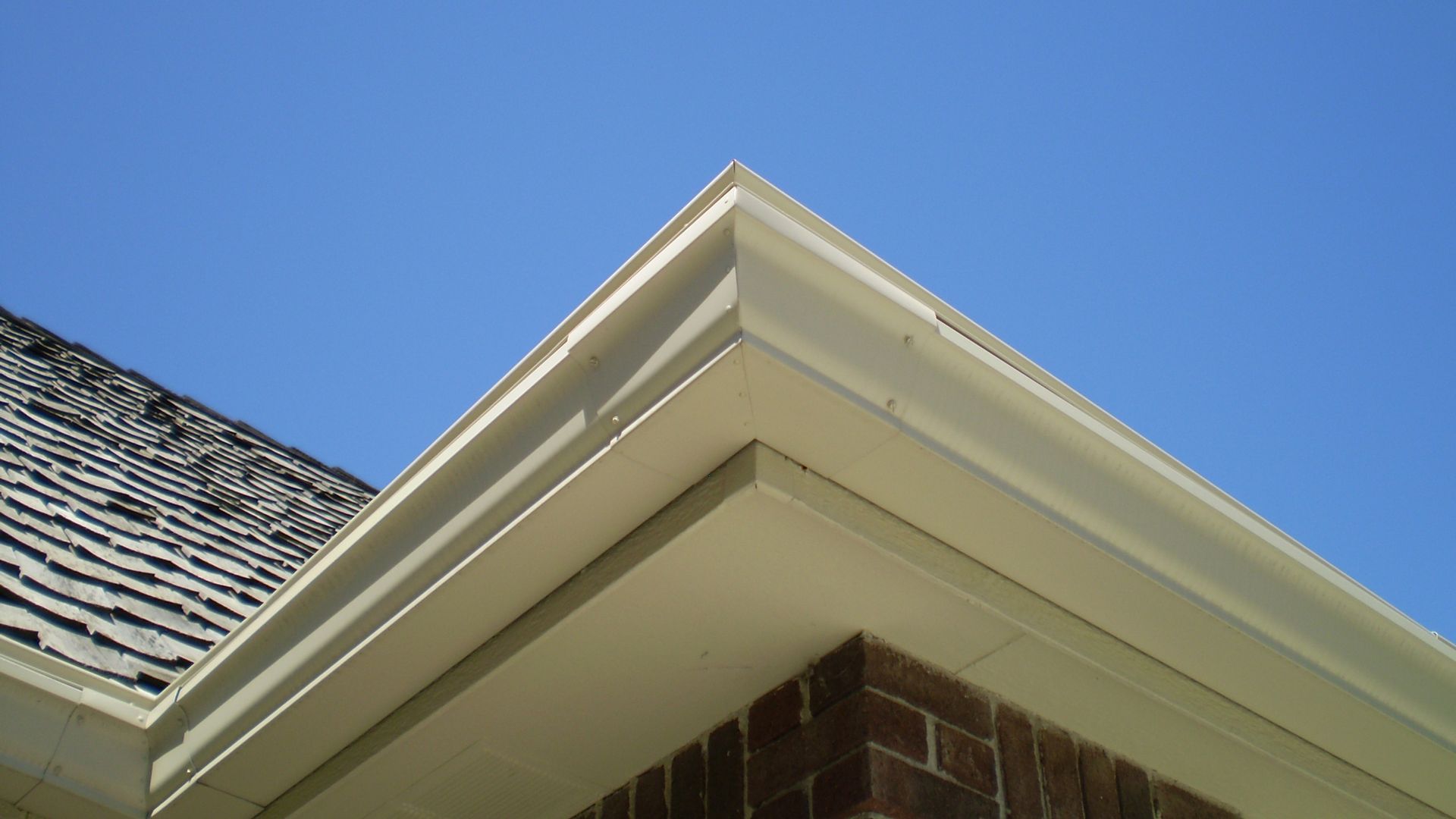 Corner of a house with white trim, gutters, brick, and dark roof tiles against a blue sky.