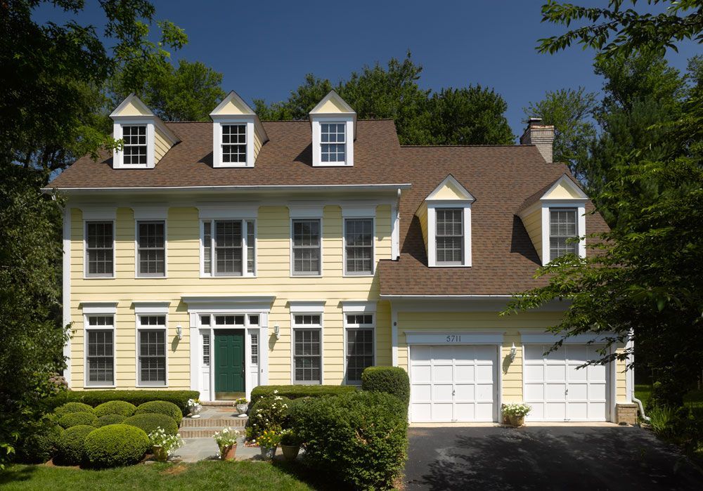 Yellow two-story house with white trim, brown roof, and three dormers. Two-car garage, green door, and landscaping.