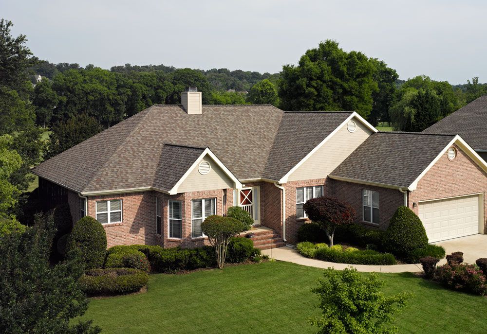 Brick house with brown roof and green lawn surrounded by trees.