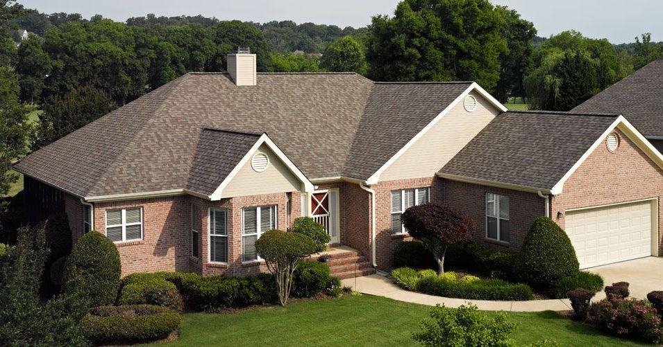 Brick house with brown shingle roof, green lawn, and shrubs; trees in the background.