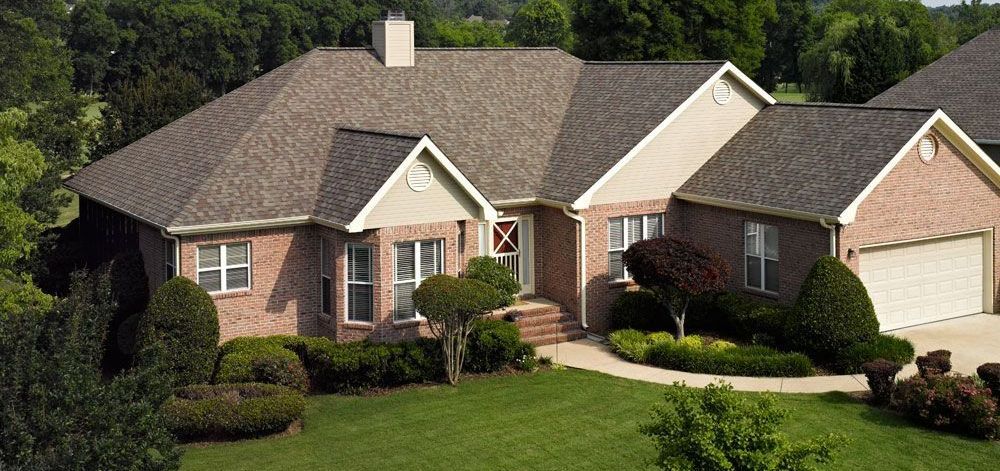 Brick house with a brown roof and a green lawn. Trees are visible in the background.