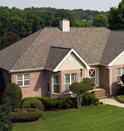 Brick house with brown shingle roof, green lawn, and surrounding trees.