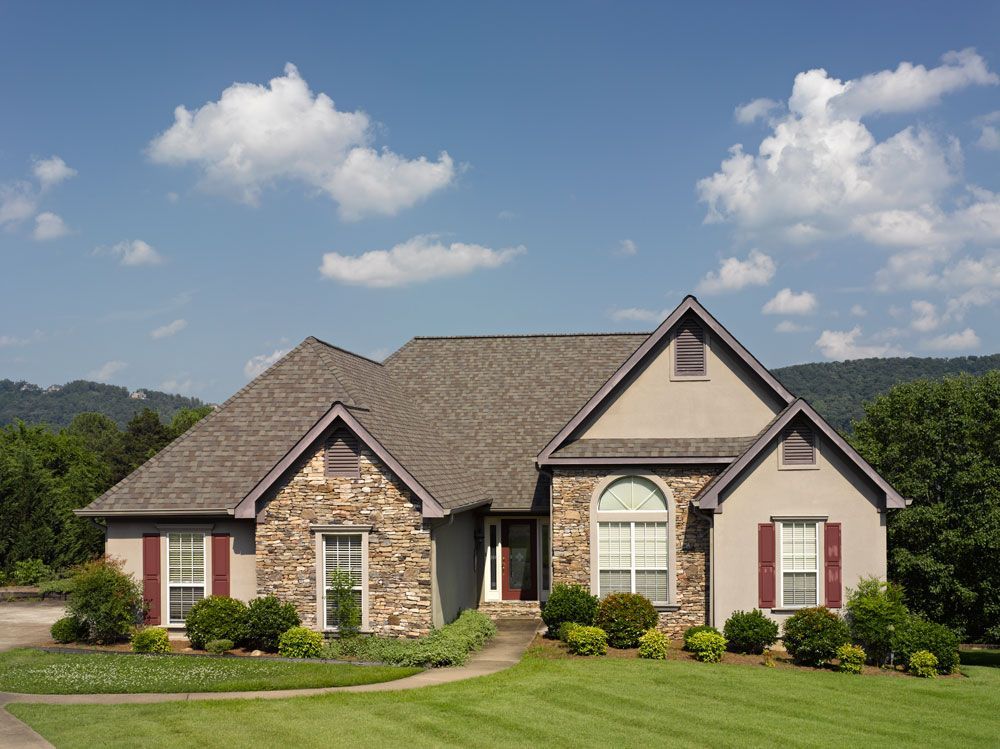 Stone and tan house with red shutters on a sunny day with green grass and trees.
