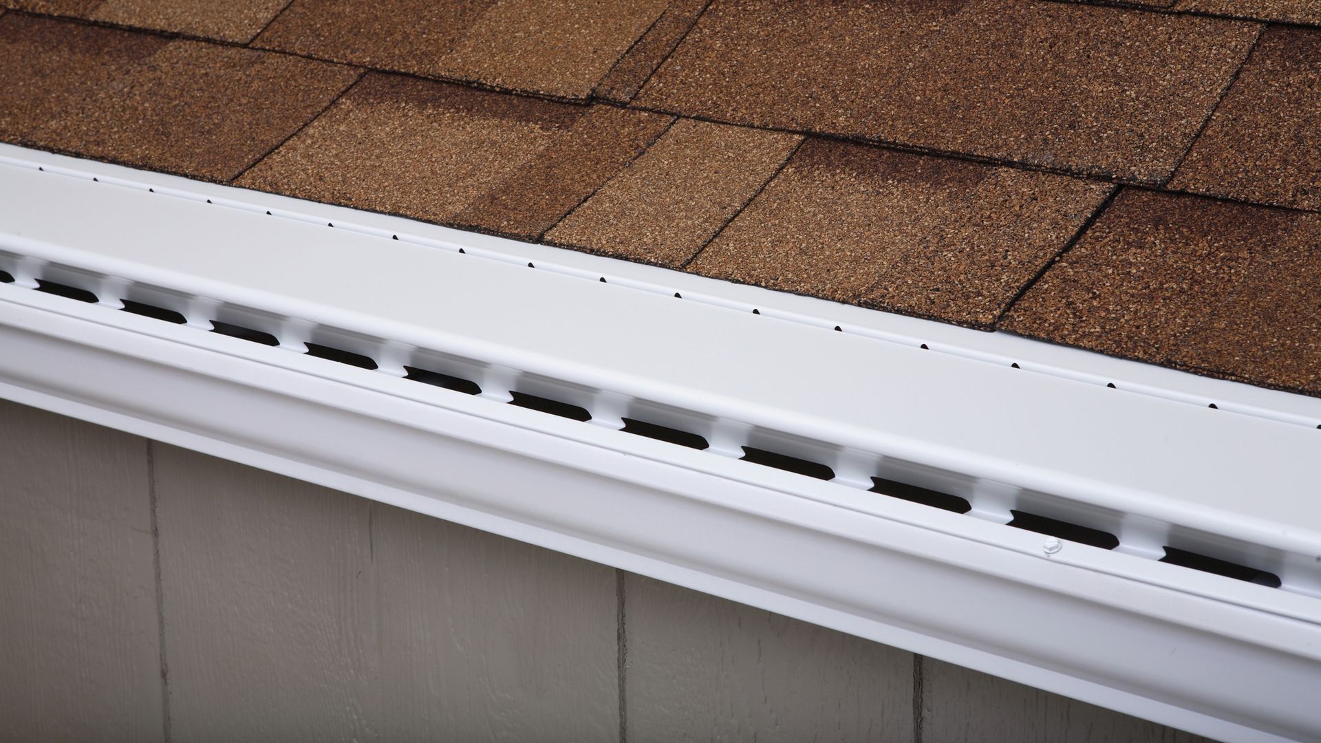 White gutter with black rectangular openings along the edge, installed below brown roof shingles and beige siding.