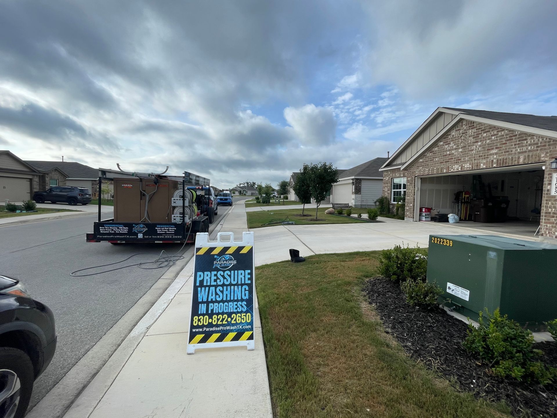 Neighborhood street with a trailer, curbside waste bin, and “pressure washing” sign near a house garage