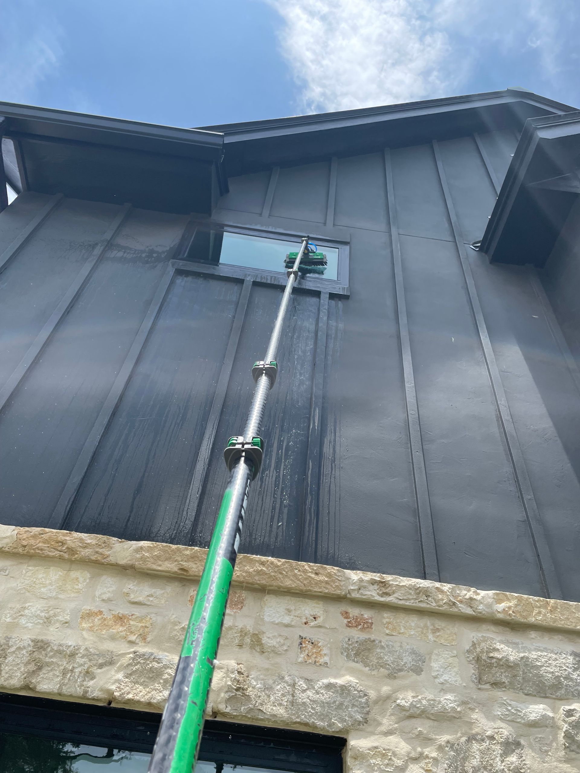 A long pole-mounted window cleaner reaches a blue window on a dark house exterior.