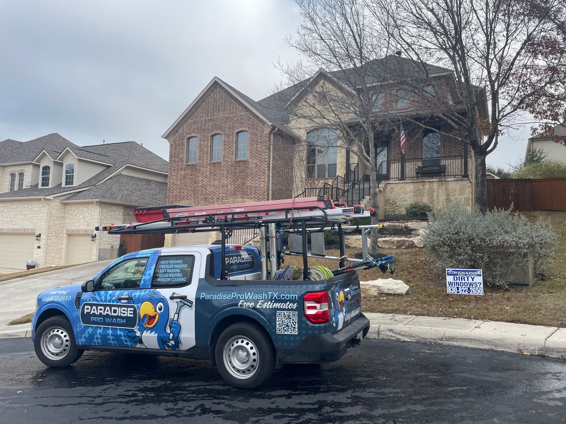 A blue HVAC service truck parked on a suburban street in front of a multi-story brick residential house.