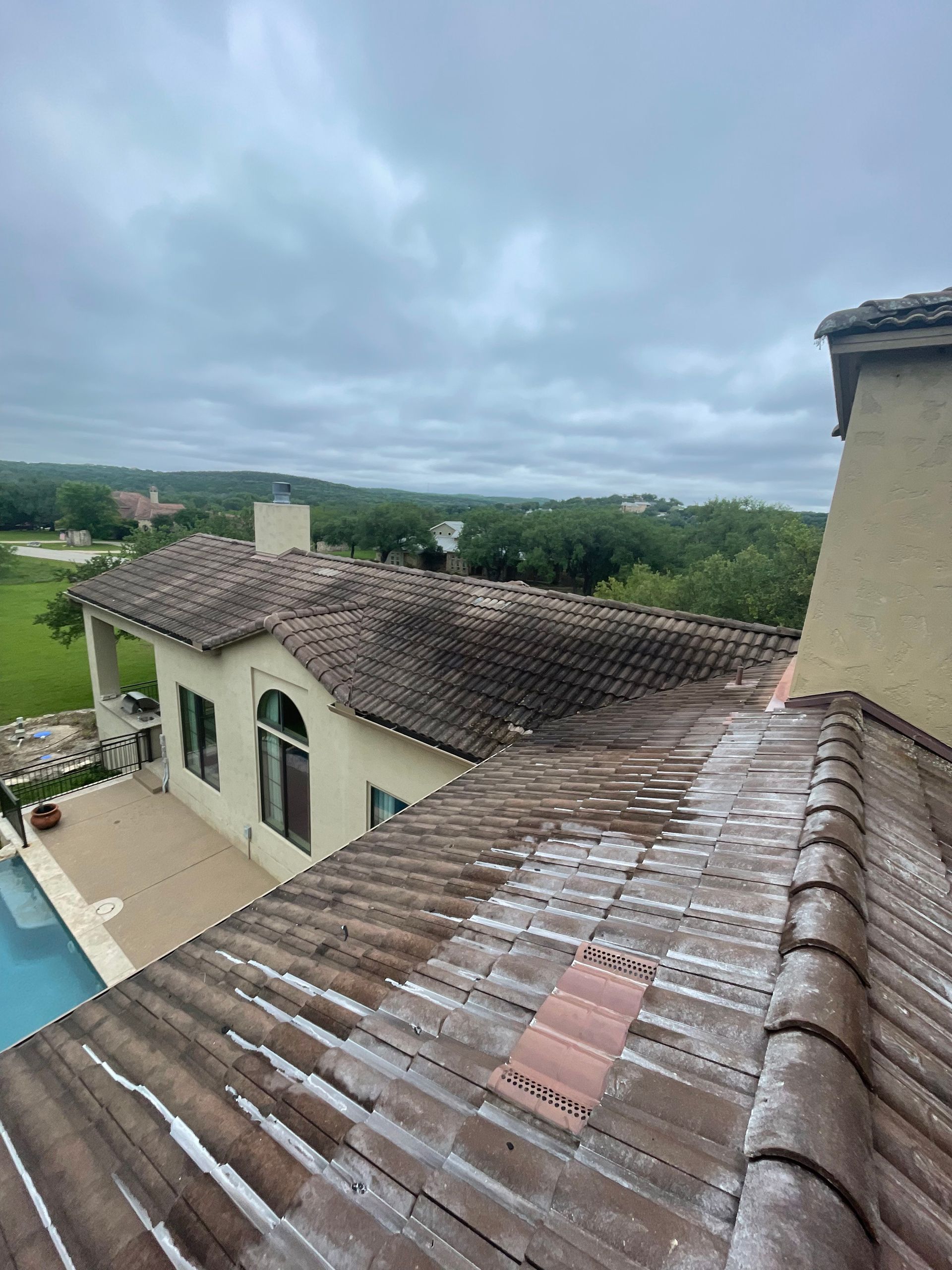 Rooftop view of a tiled villa beside a pool, with green fields and cloudy sky beyond