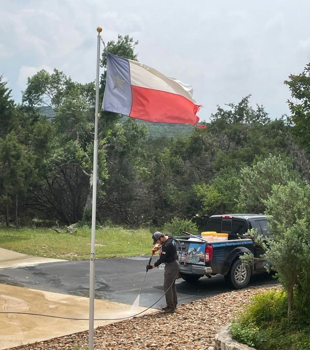 Person pressure washing a driveway next to a Texas flag and truck. Trees in background.