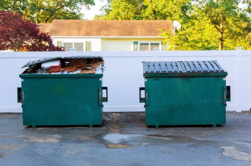 Two green dumpsters are sitting next to each other in front of a white fence.