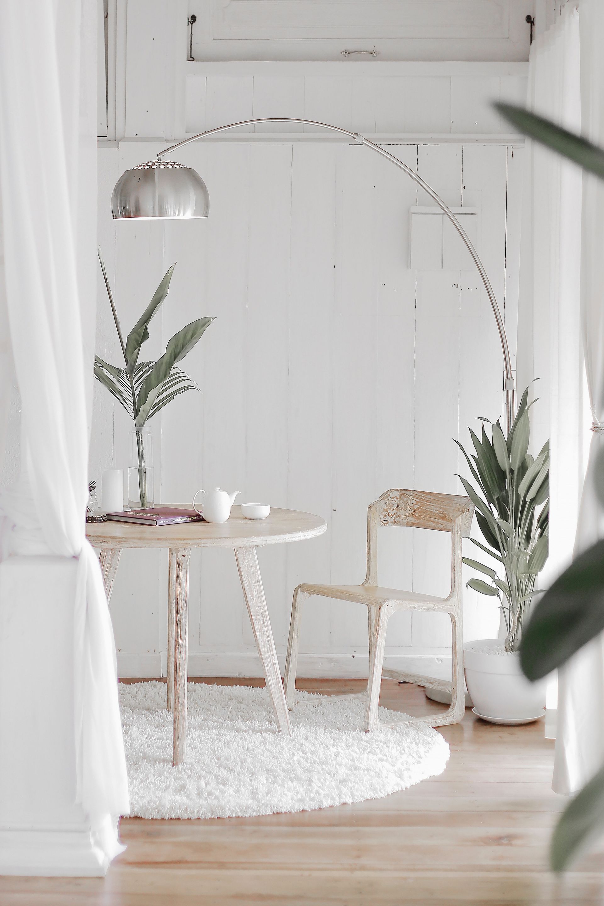 Eine minimalistische, weiße Essecke mit einem runden Tisch, einem Holzstuhl, einer hohen Bogenstehlampe und zwei Topfpflanzen.