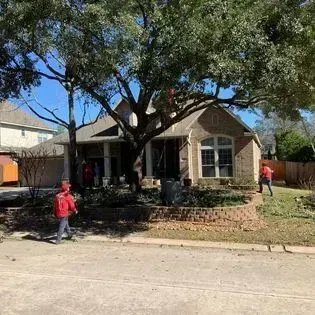 Single-story brick house with large trees and two people in red shirts outside by the sidewalk.
