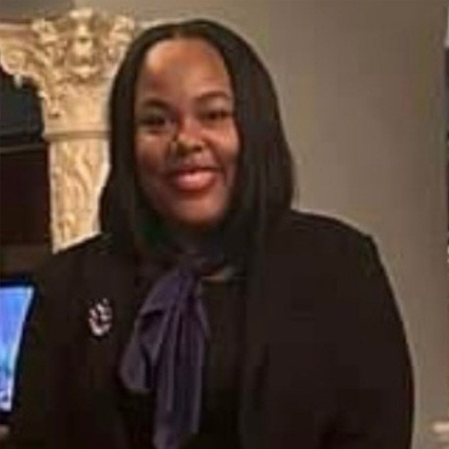 Woman in a black blazer and purple bow blouse, smiling indoors near a decorative column.