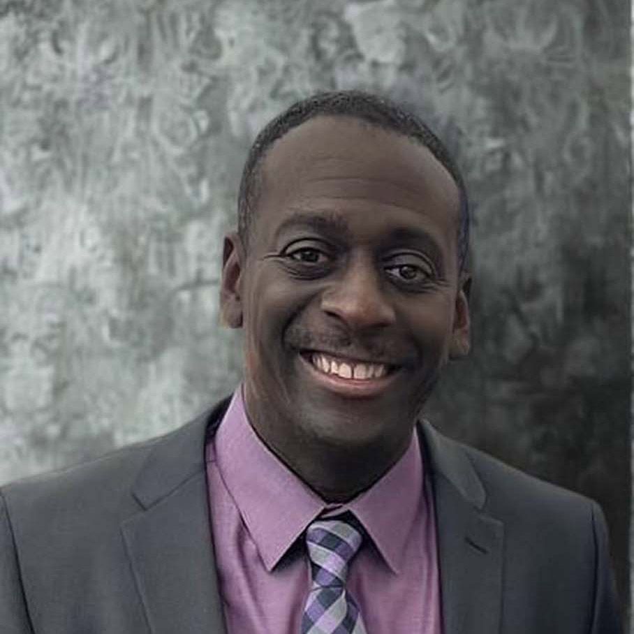 Smiling man in a gray suit and purple shirt against a textured gray background