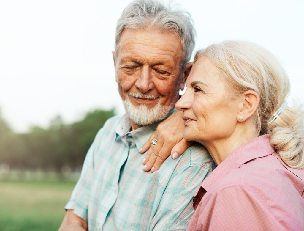 Smiling elderly couple embracing outdoors in a green park, with soft sunlight and blurred trees in the background