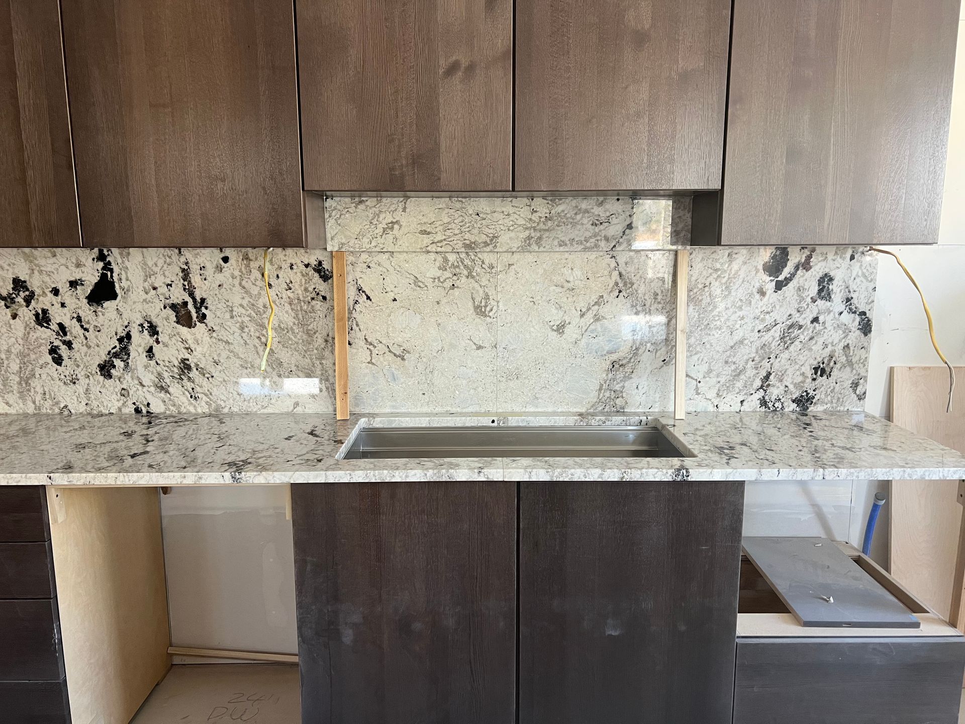 Modern kitchen with dark wood cabinets, speckled stone backsplash, and white countertop under construction