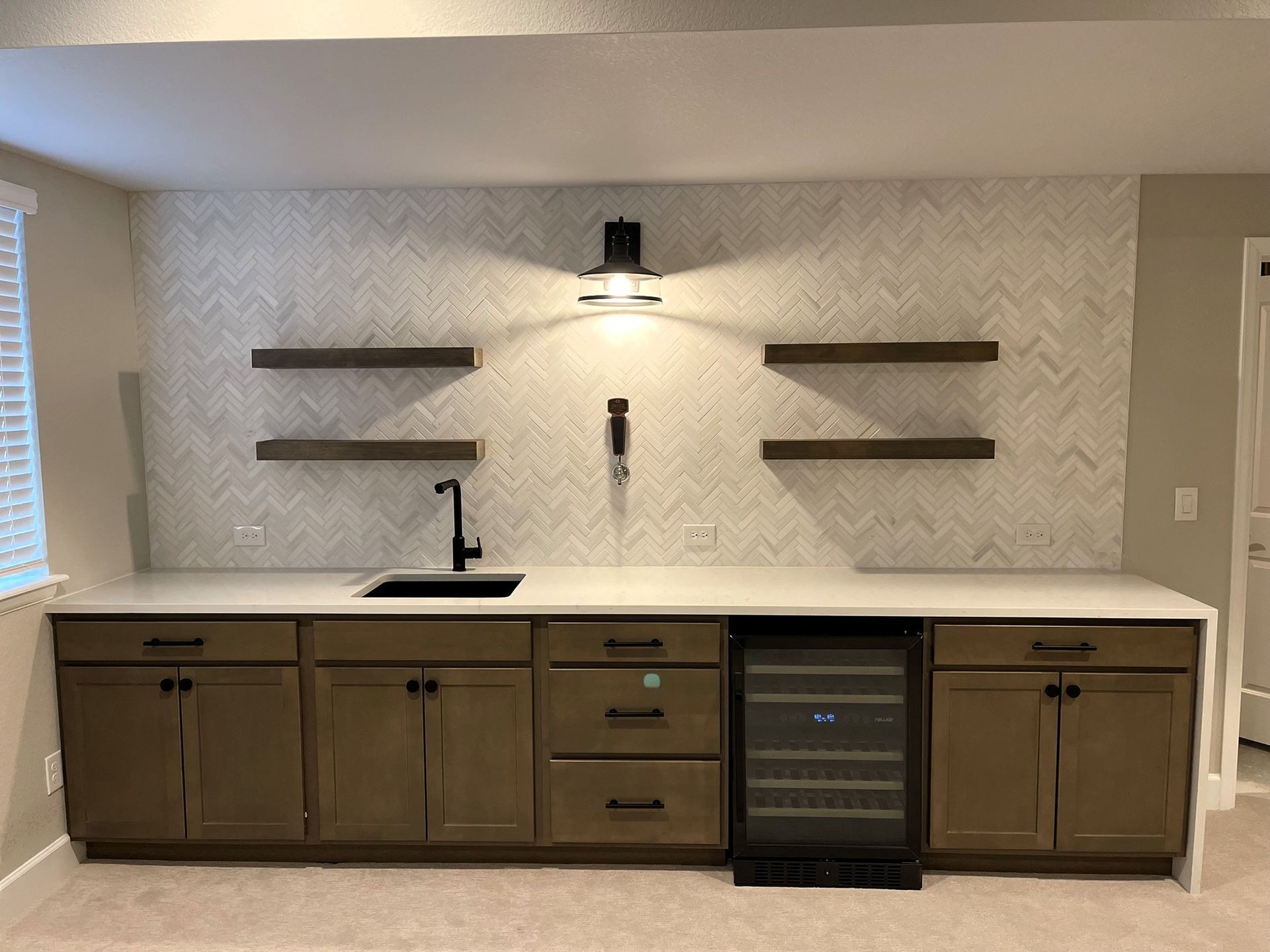 Basement wet bar with dark cabinets, white countertop, black sink, wine cooler, and floating shelves.