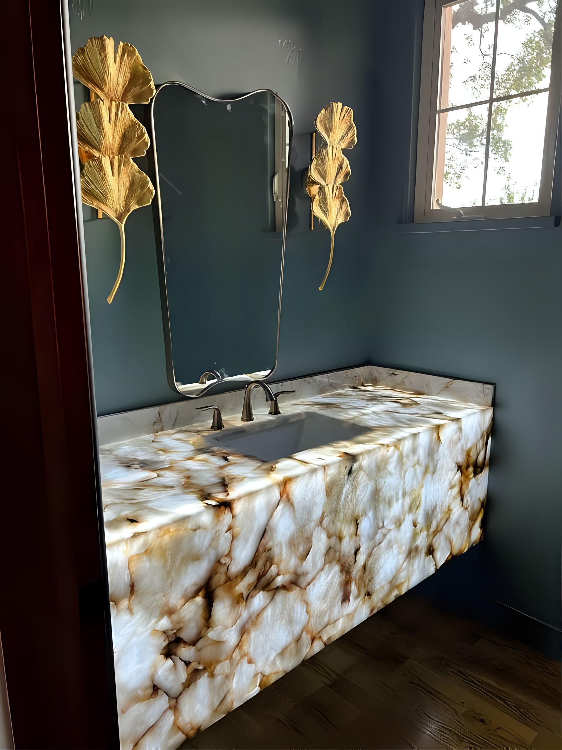 Marble vanity with mirror in a dark teal bathroom, accented by gold leaf wall decor and a window.