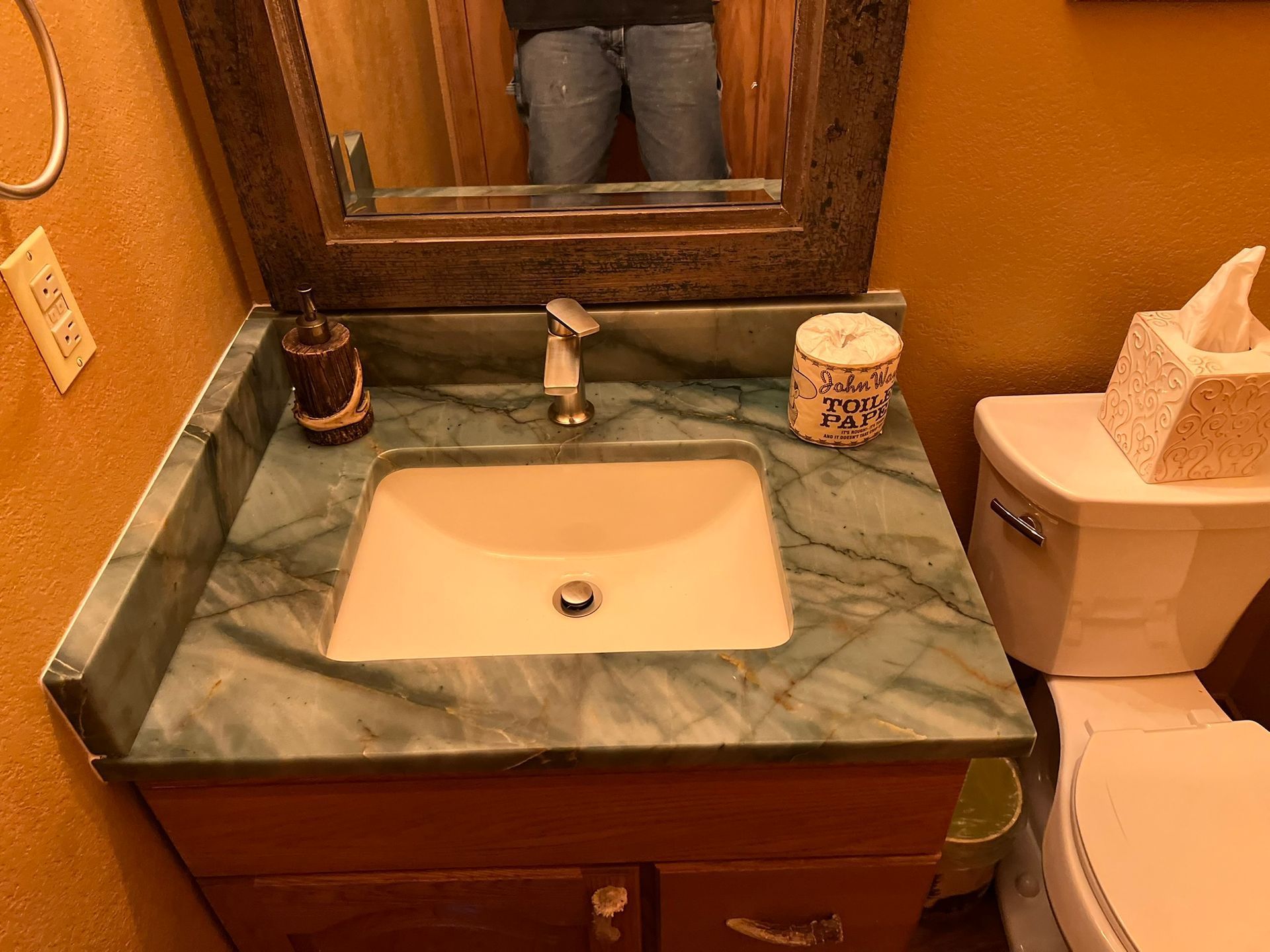 Bathroom sink with green marble countertop, mirror, soap dispenser, and toilet in an orange-painted room