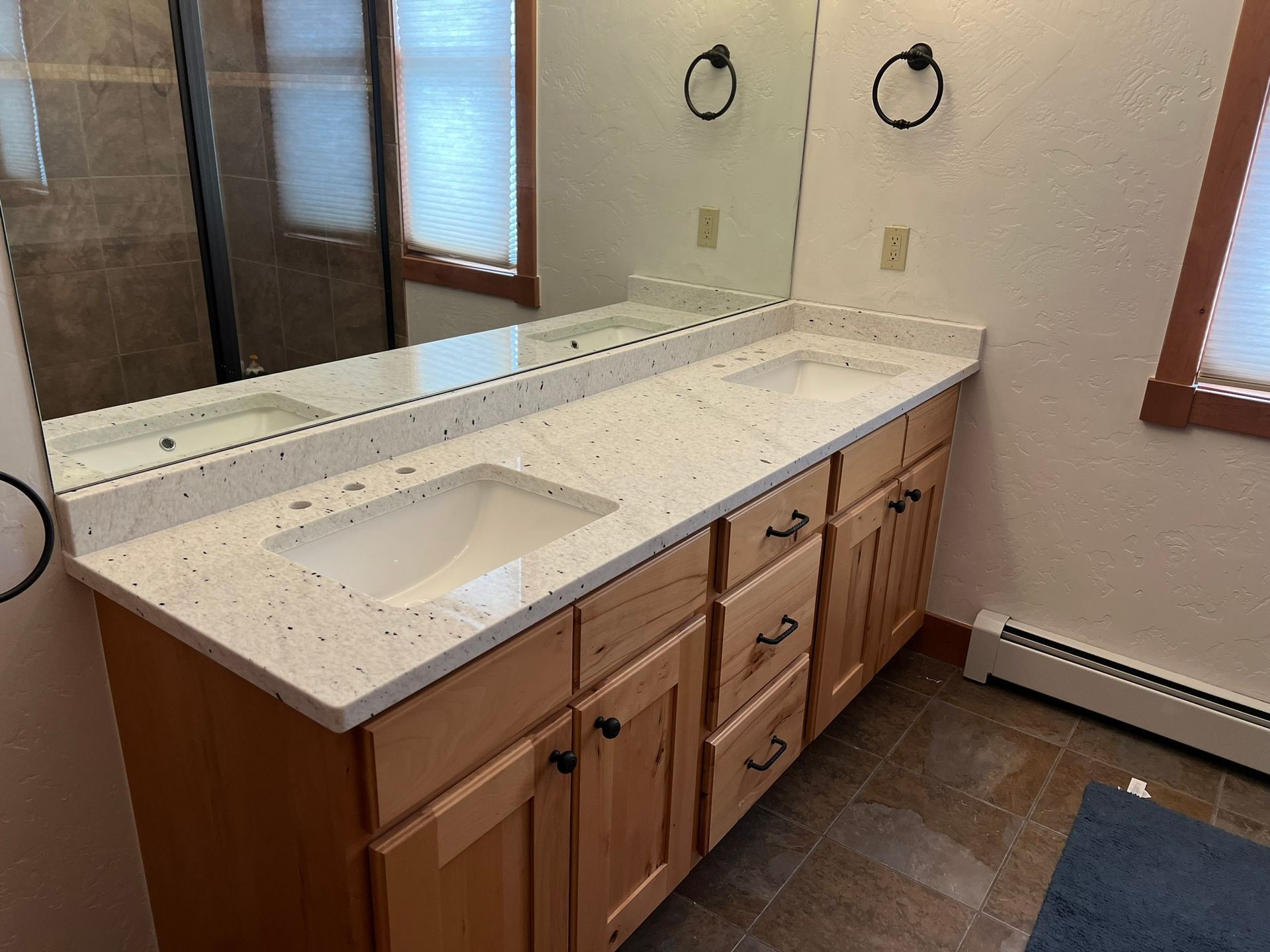 Bathroom vanity with speckled countertop, rectangular sink, wood cabinets, and large mirror by a window.