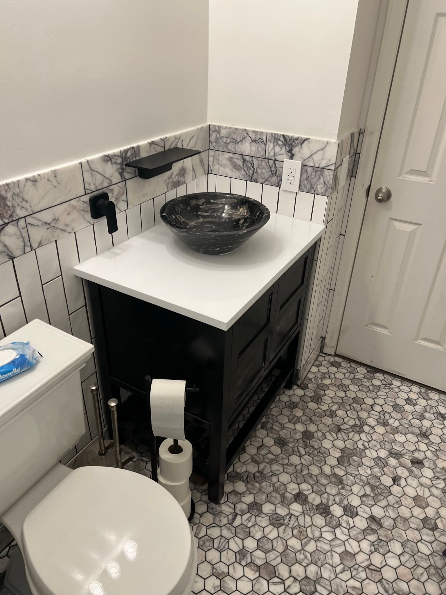 Small bathroom with patterned tile, black vanity, vessel sink, toilet, and white door.