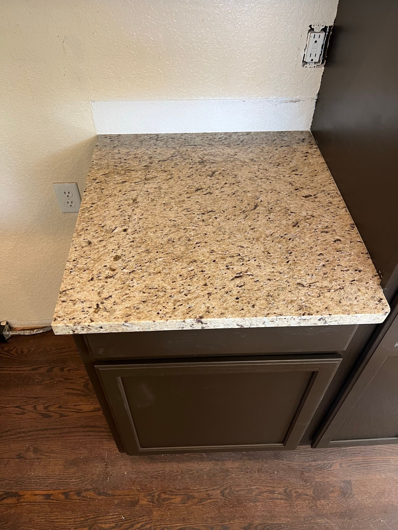 Small speckled countertop above a dark cabinet in a corner by a wall and wood floor.