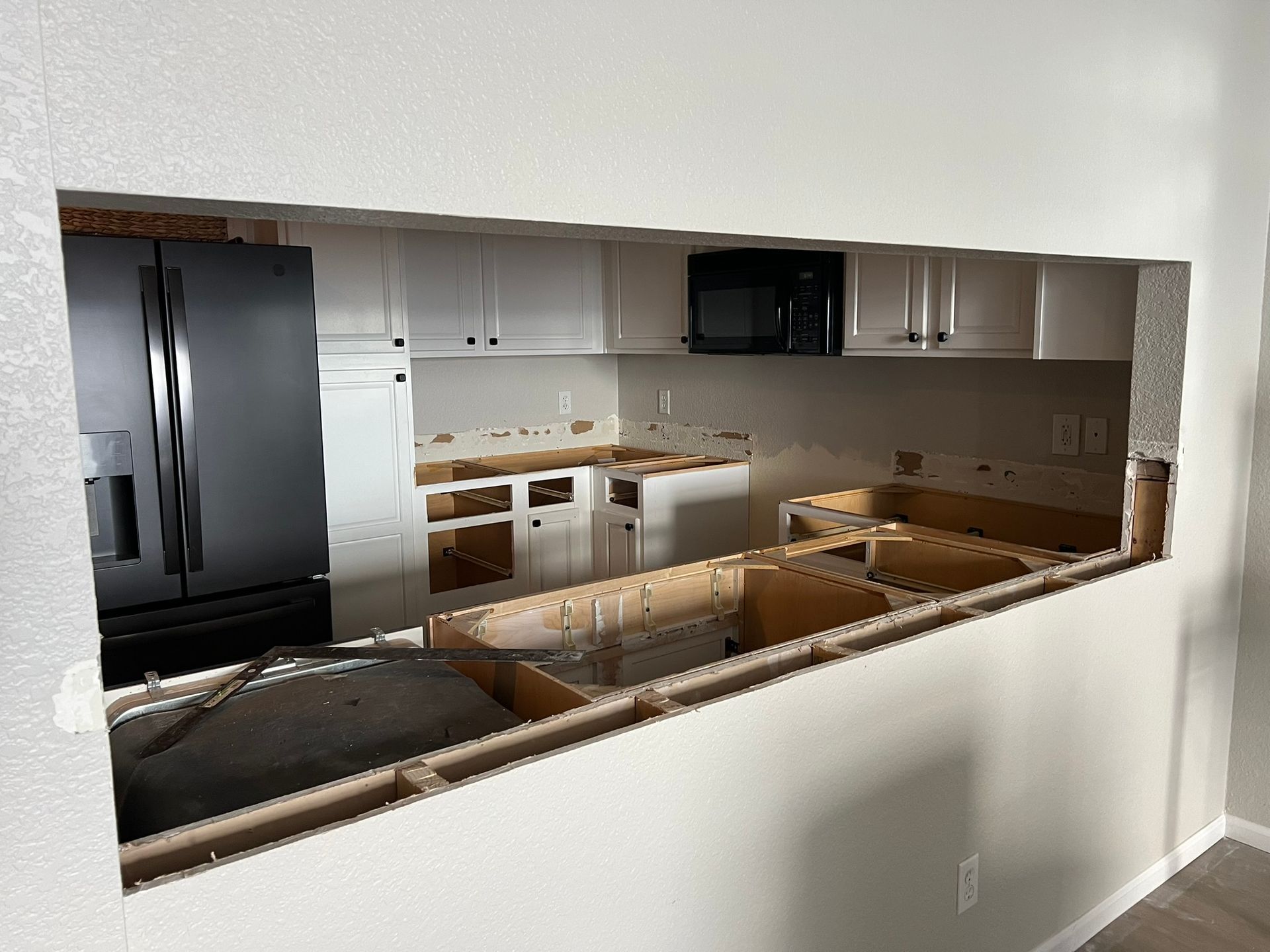 Kitchen viewed through a large wall opening, showing black appliances, white cabinets, and unfinished countertops
