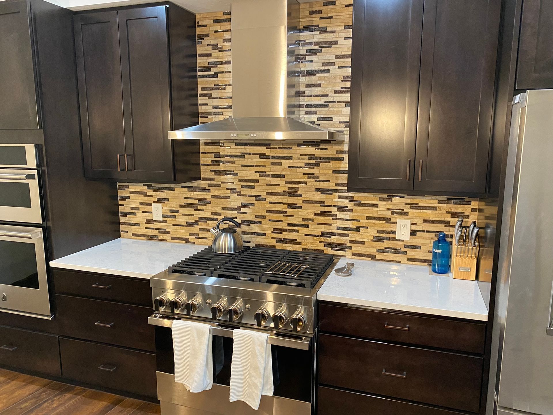 Modern kitchen with dark wood cabinets, stainless steel stove and hood, white countertops, and mosaic tile backsplash