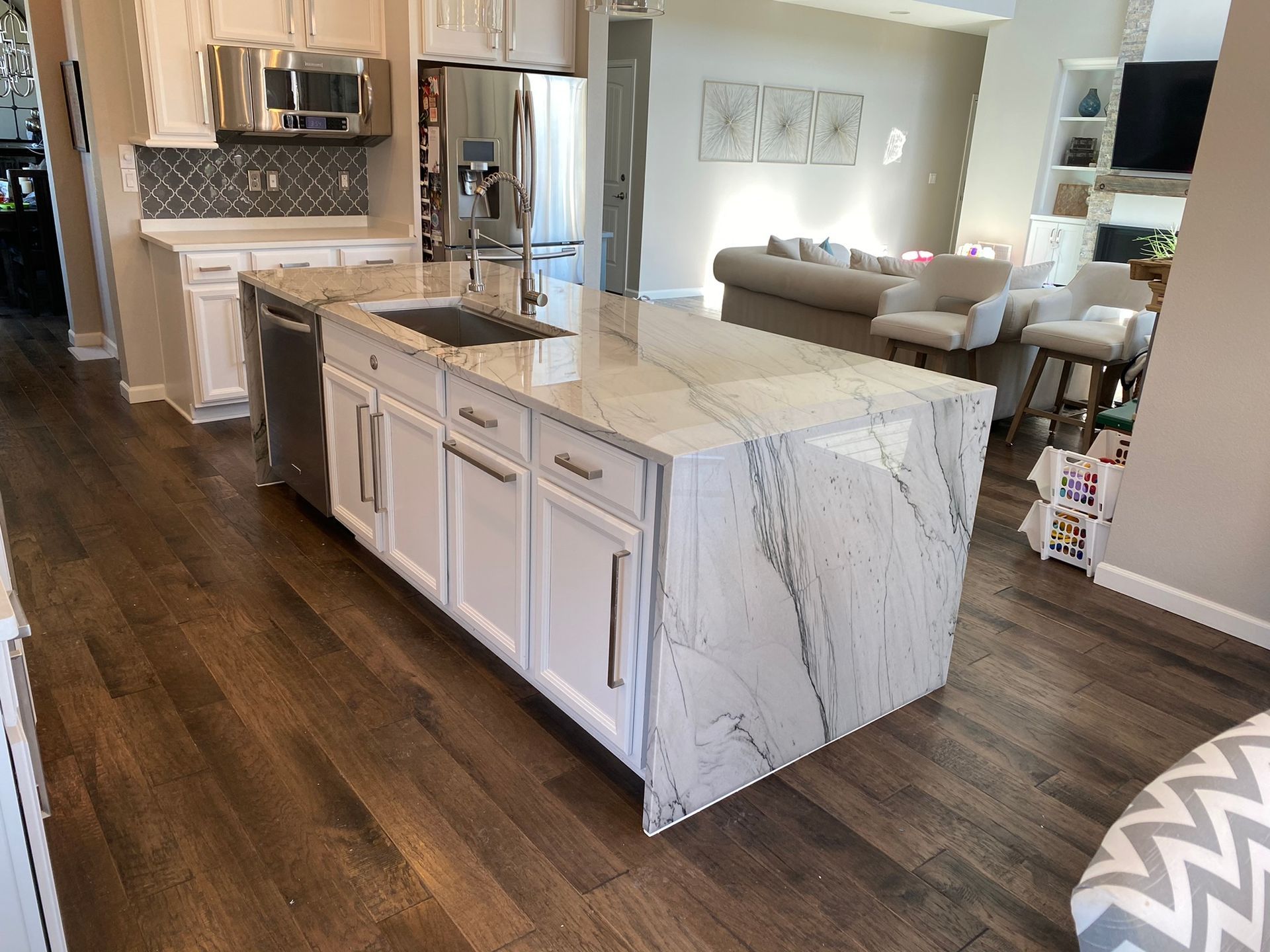 Bright kitchen with marble island, white cabinets, dark wood floors, and a living area in the background