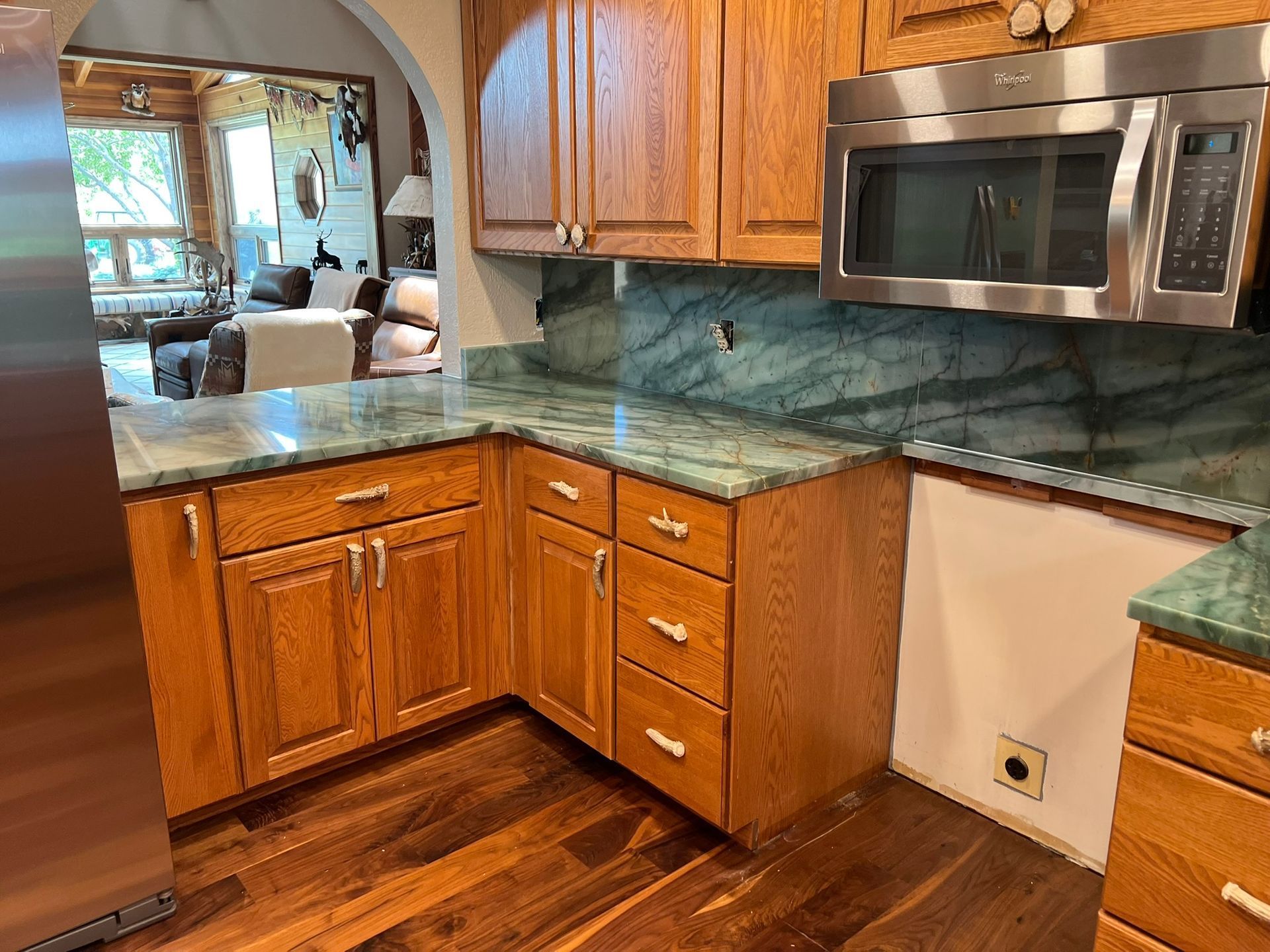 Wooden kitchen with green granite countertops, stainless microwave, and a pass-through opening to a living room.