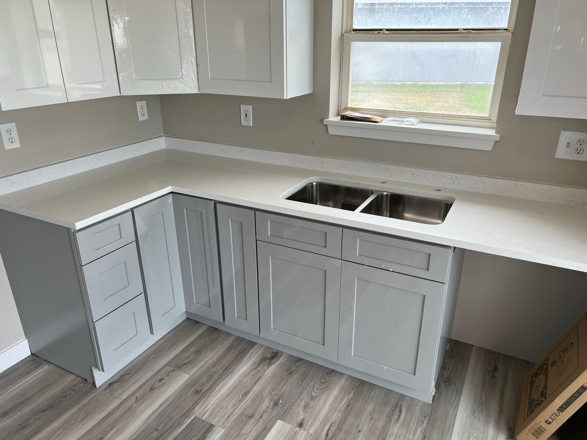 New white kitchen cabinets and countertops with a double sink and gray wood-look flooring.