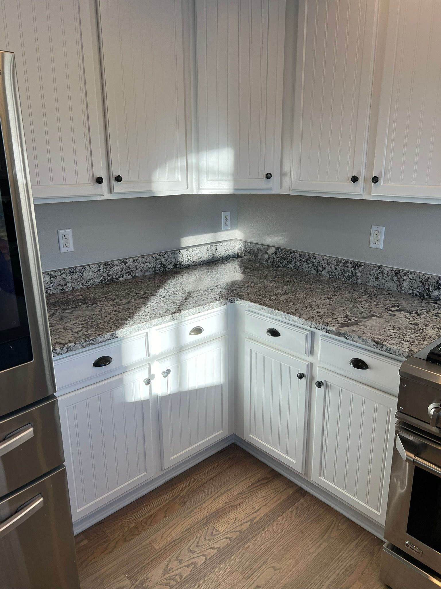 Empty kitchen corner with white cabinets, speckled granite countertops, stainless steel appliances, and wood floor.