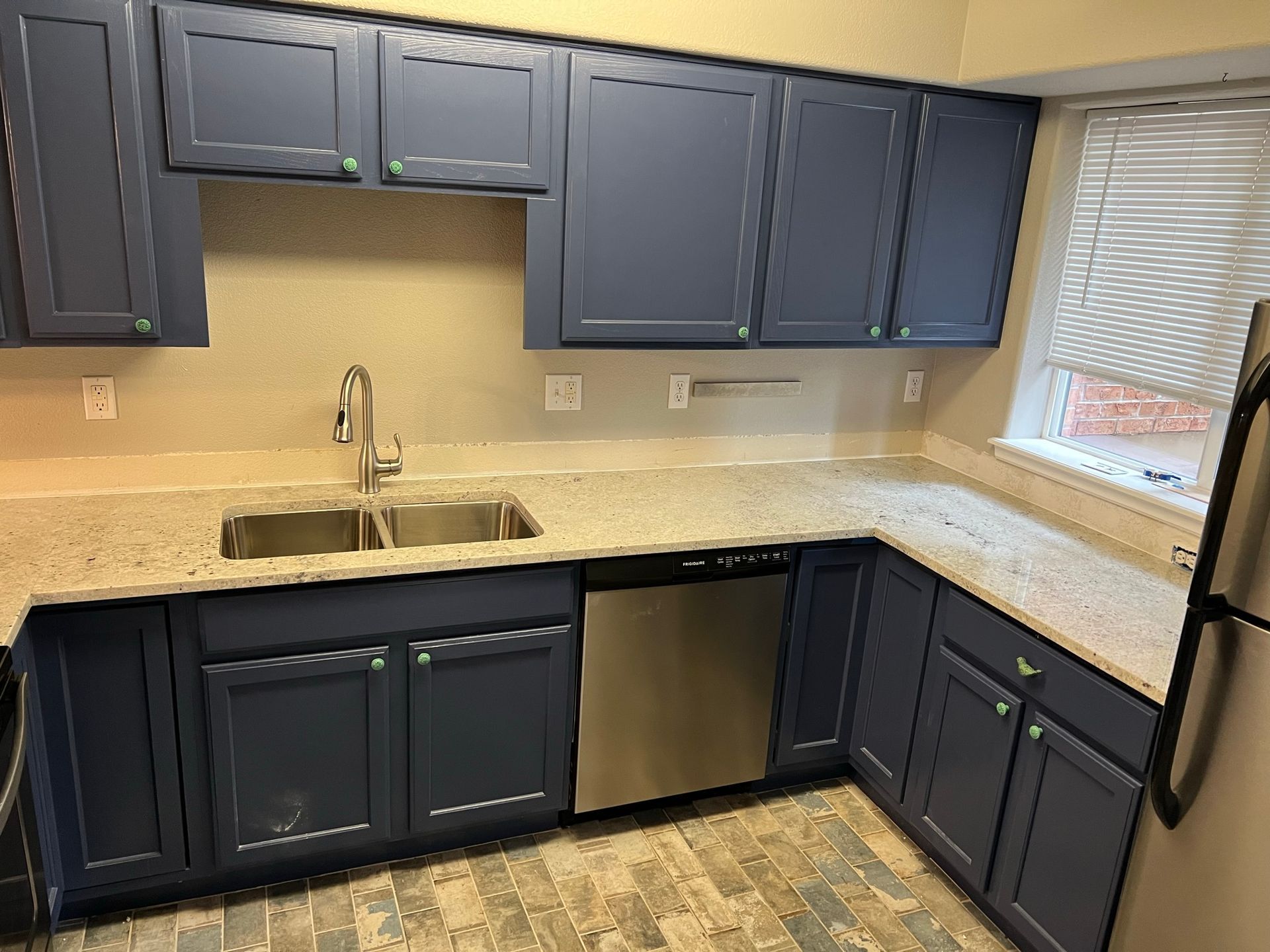 Empty kitchen with dark blue cabinets, light countertops, stainless sink, and a window with blinds.
