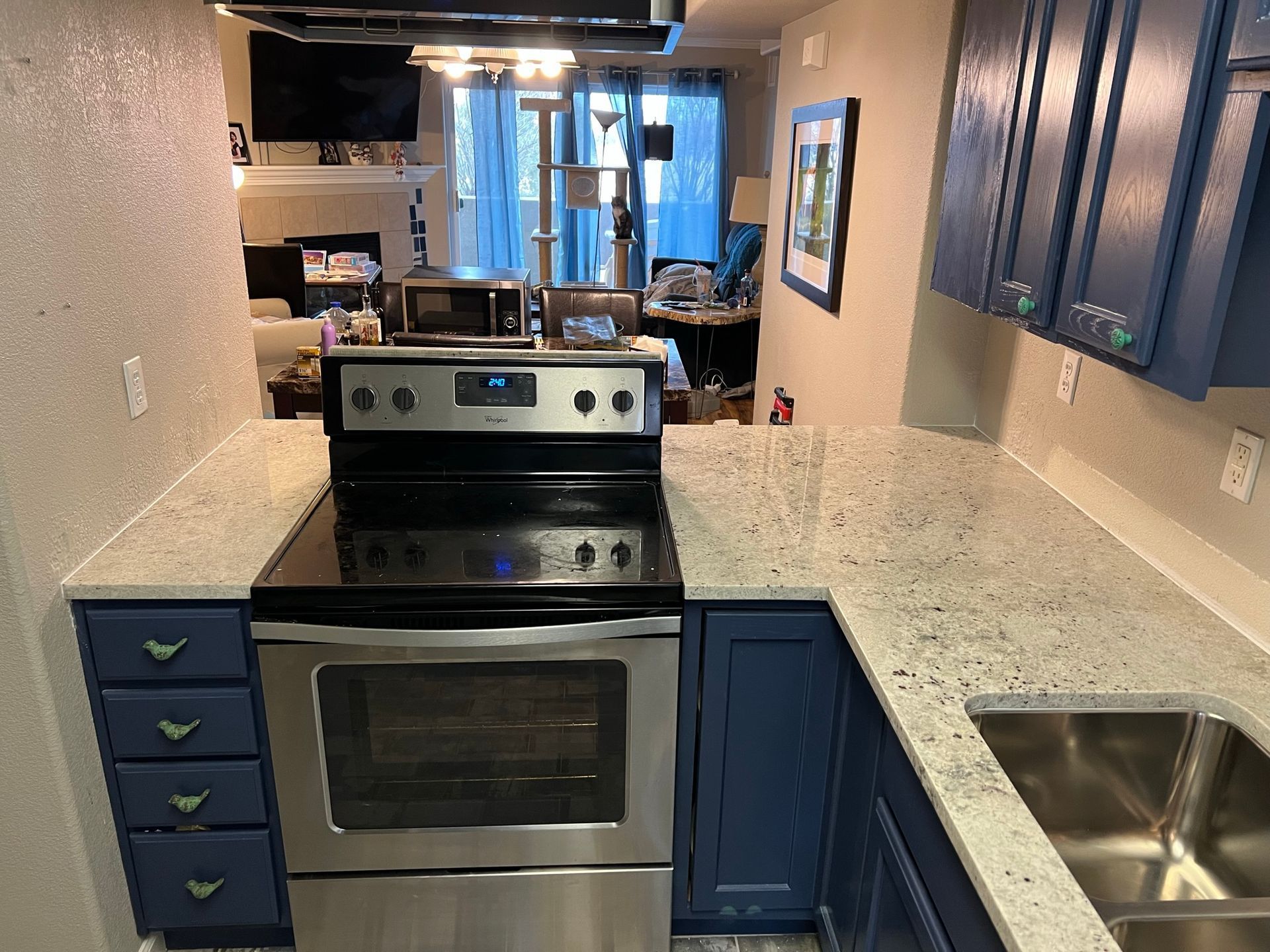 Compact kitchen with blue cabinets, stainless stove, speckled counters, and a sink under a window.