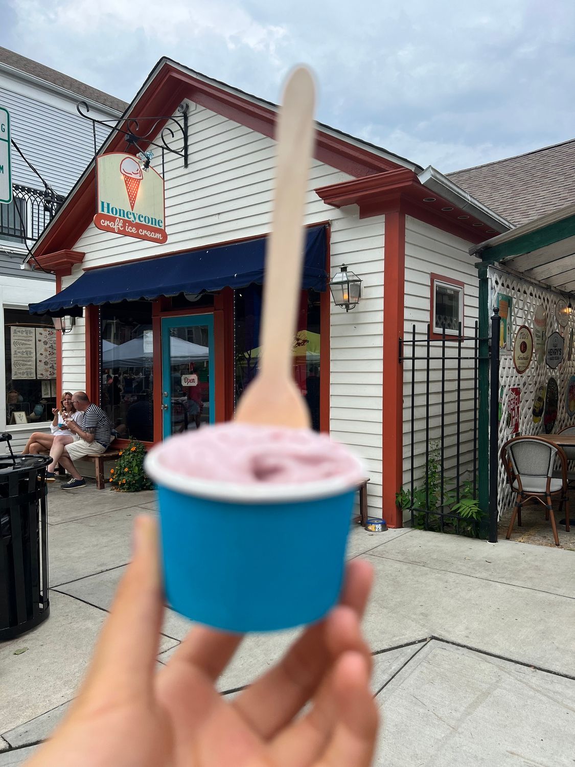 A person is holding a cup of ice cream with a wooden spoon in front of a building.
