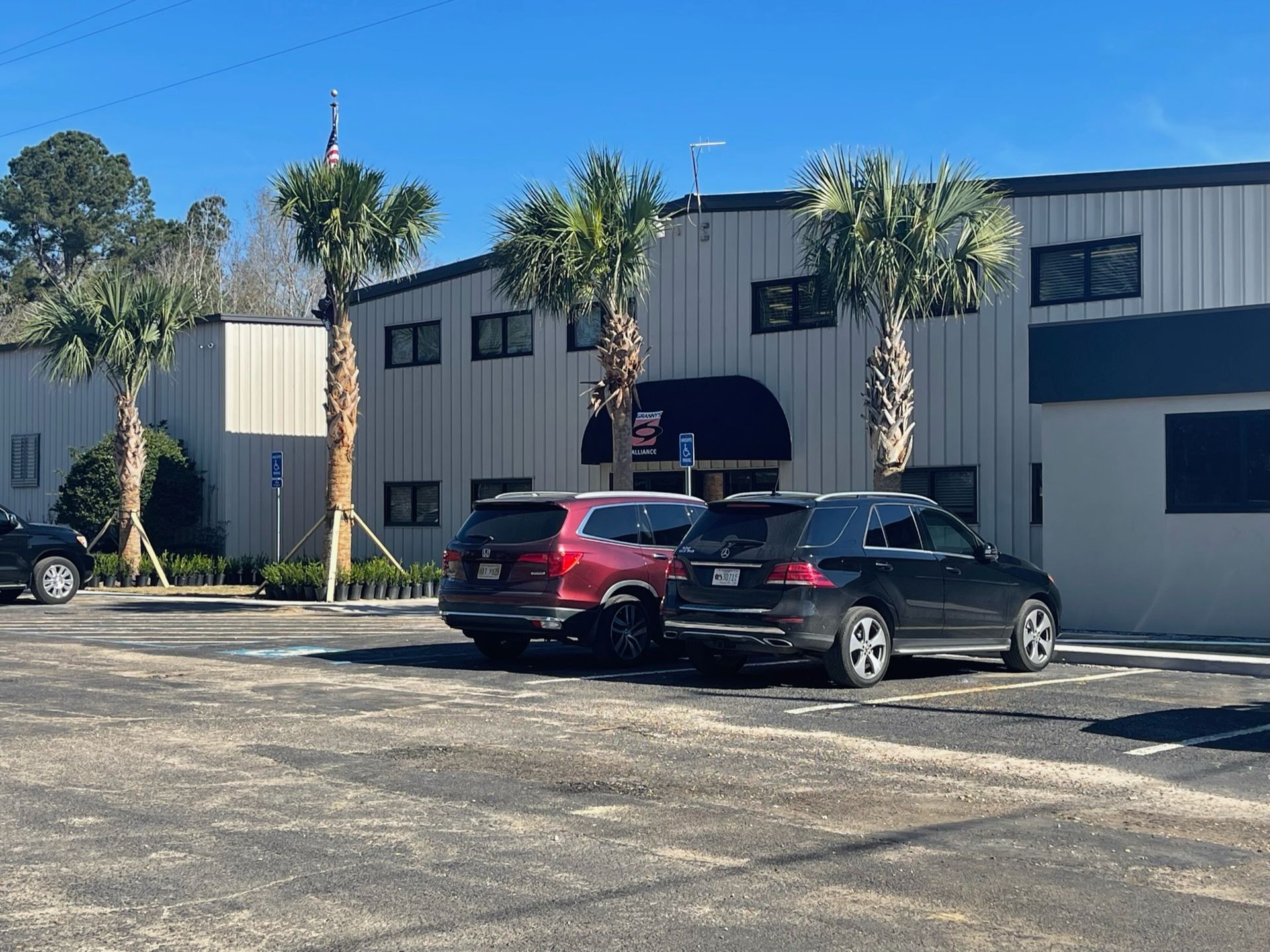 Building with metal siding, palm trees, parked cars, and paved parking lot under a blue sky.