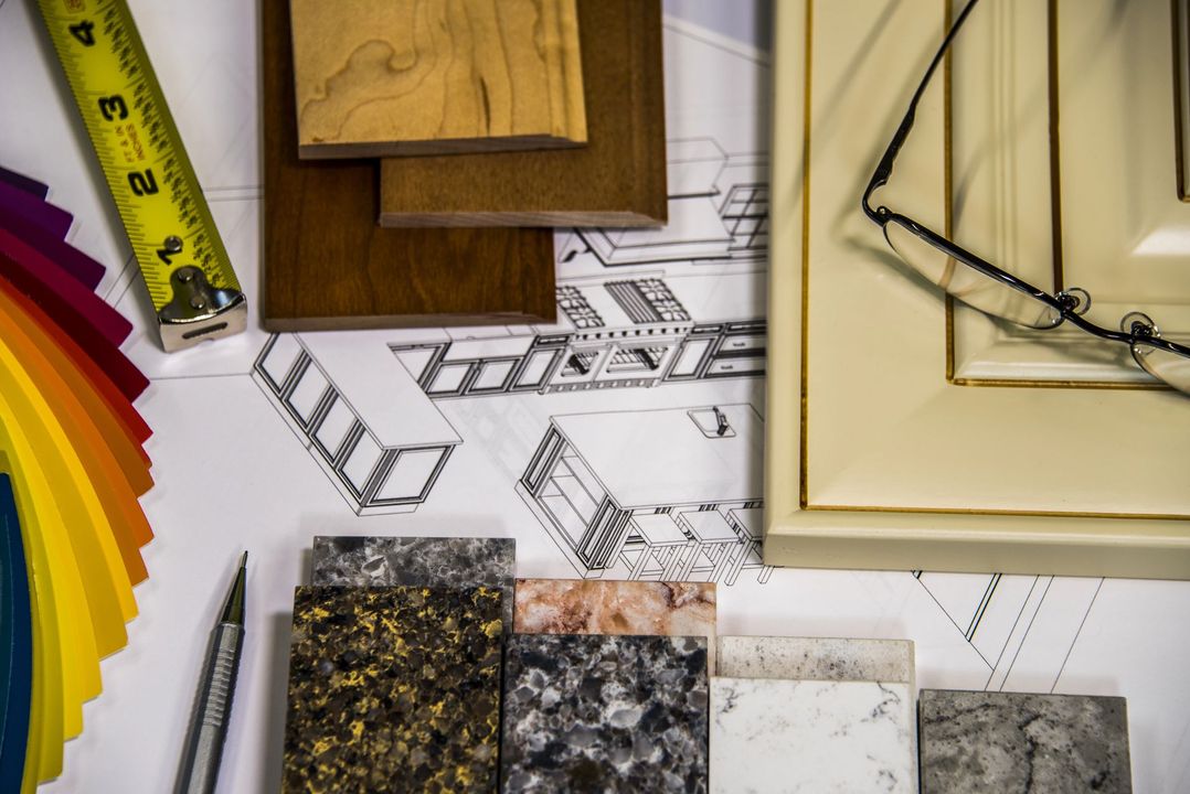 Materiali e strumenti di progettazione disposti su planimetrie architettoniche, con legno, pietra, campioni di vernice e un metro a nastro.