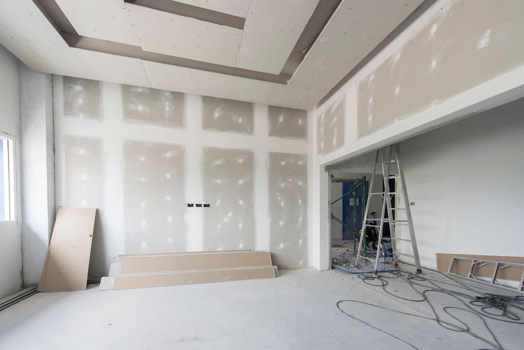 Stanza non rifinita con riparazioni al muro a secco, scala e materiali edili su un pavimento bianco.