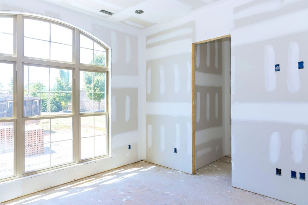 Luminosa stanza non rifinita con grande finestra, pareti in cartongesso bianco e luce naturale che entra