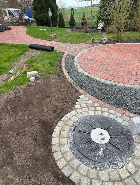 Giardino paesaggistico con patio in mattoni, sentiero di ghiaia, prato e focolare circolare in pietra in primo piano.