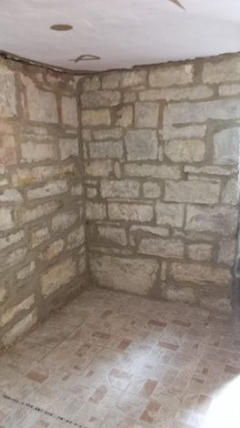 Stanza vuota con pareti in pietra, pavimento piastrellato e angolo del soffitto non rifinito.