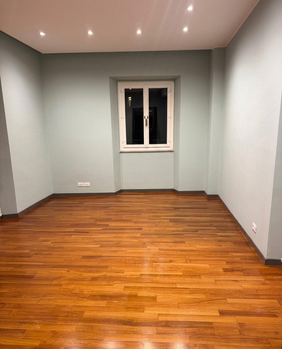 Stanza vuota con pavimento in legno, pareti grigie, faretti a incasso sul soffitto e una piccola finestra centrata sulla parete di fondo.