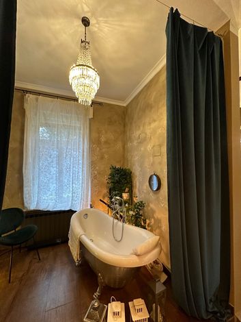 Elegante bagno con pareti dorate, lampadario di cristallo, vasca con piedini e tende scure.