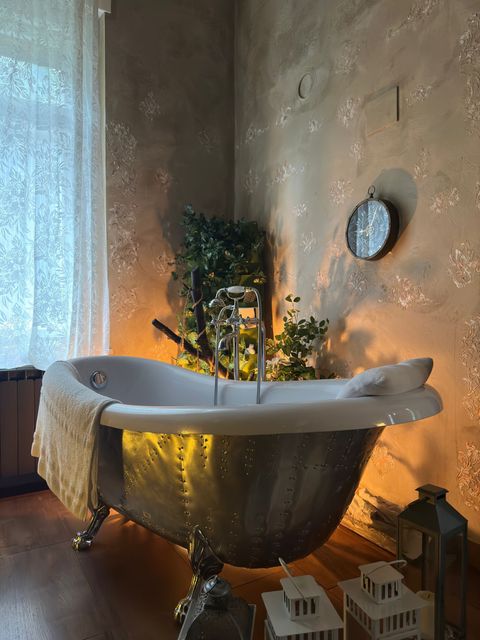 Accogliente vasca da bagno con piedini, candele, luci decorative, pianta e lanterne in un bagno illuminato da una luce soffusa.