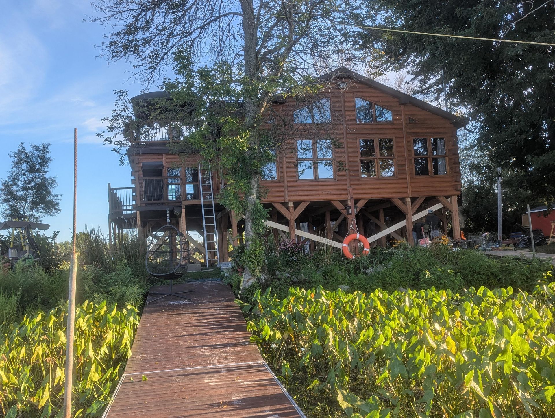 Une maison sur pilotis en bois surplombant une zone marécageuse, accessible par une longue passerelle en bois sous un ciel bleu azur.
