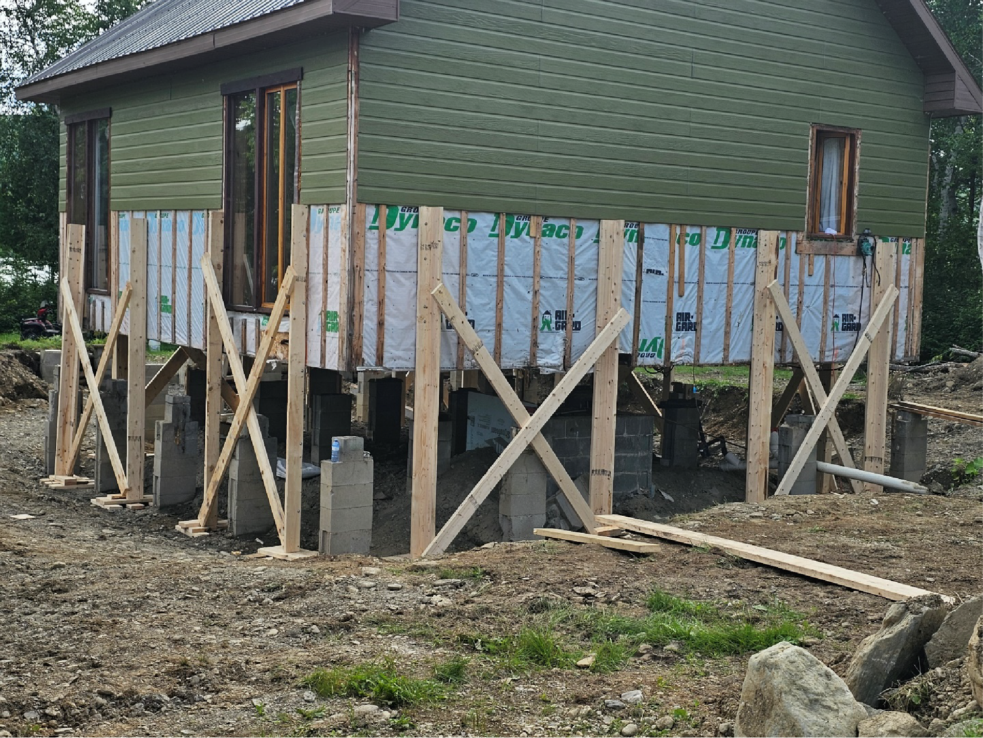 Une petite maison au bardage vert, soutenue par des pilotis en bois et des blocs de béton provisoires, en cours de rénovation.