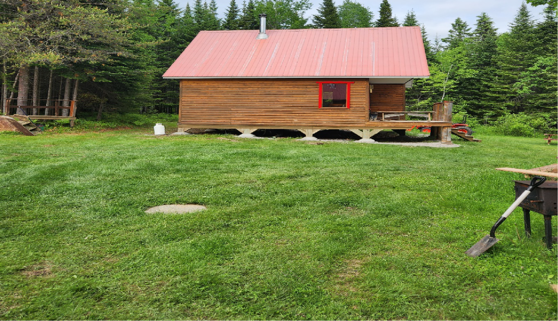 Cabane en bois avec toit et fenêtre rouges, située dans un champ herbeux, arbres en arrière-plan.