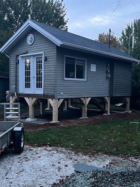 Cabane grise avec portes et fenêtre blanches, construite sur pilotis. Allée de gravier et caravane au premier plan.
